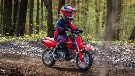 Jeune enfant souriant assis sur une Honda CRF50F rouge, dans un environnement extérieur adapté à la moto tout-terrain.