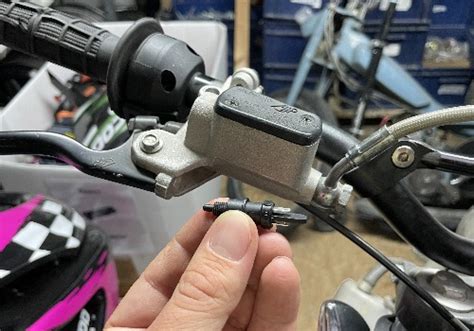 Vue éclatée d'un contacteur de frein avant de moto, montrant les lamelles métalliques, le ressort et les rivets en plastique.